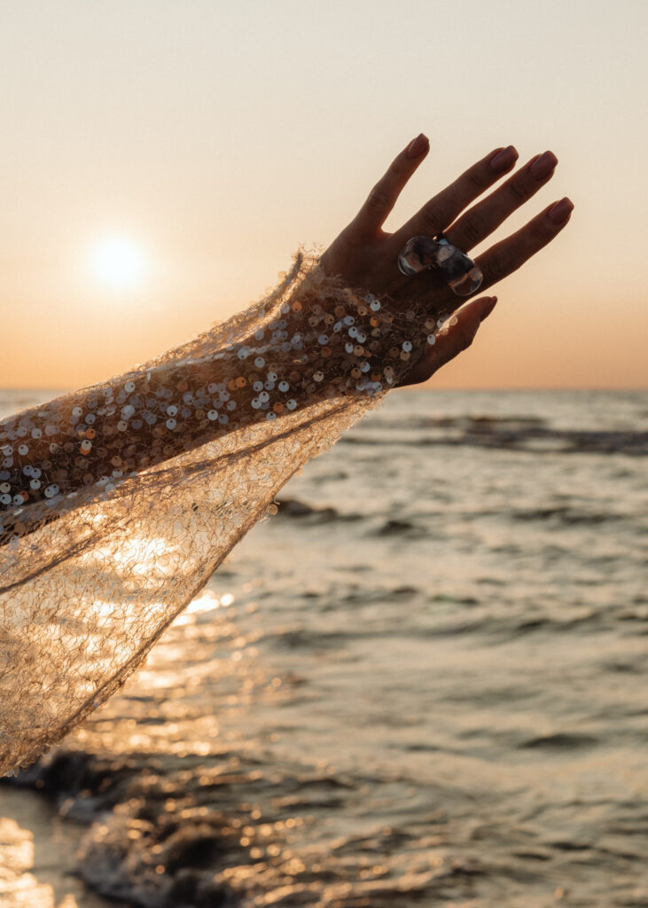 Hand reaching toward the sunlight over the ocean, adorned in a sequin-covered sleeve, symbolizing hope and light.